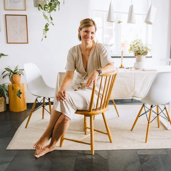 Lächelnde Frau sitzt in einem gemütlichen, nachhaltig gestalteten Essbereich mit Holzmöbeln und Zimmerpflanzen, der minimalistisches und umweltfreundliches Innendesign zeigt.