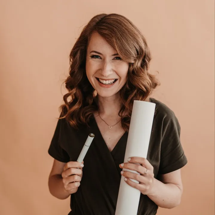 Porträt einer lächelnden Frau mit lockigem Haar namens Eva Hofmann von Recreative Interior, die eine Rolle Papier und einen Stift hält, Partnerin eines Startups für nachhaltige Farben namens Mynt aus Deutschland, vor einem beigefarbenen Hintergrund.